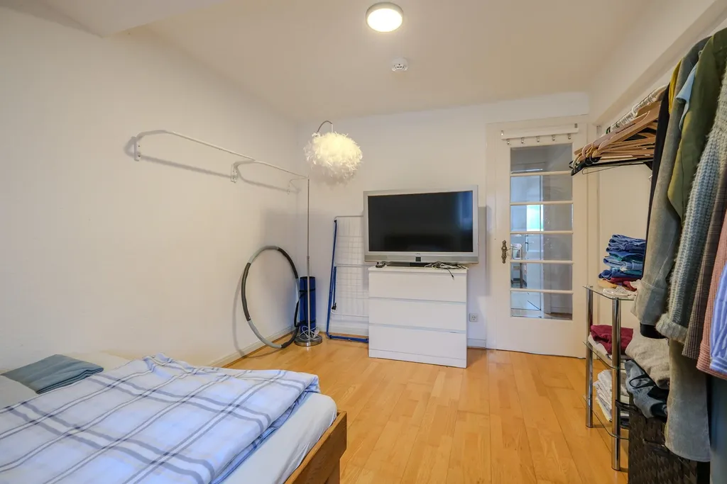 Bedroom with wooden floor, a bed with blue checkered bedding, a television on a white dresser, and clothing on an open clothes rack.