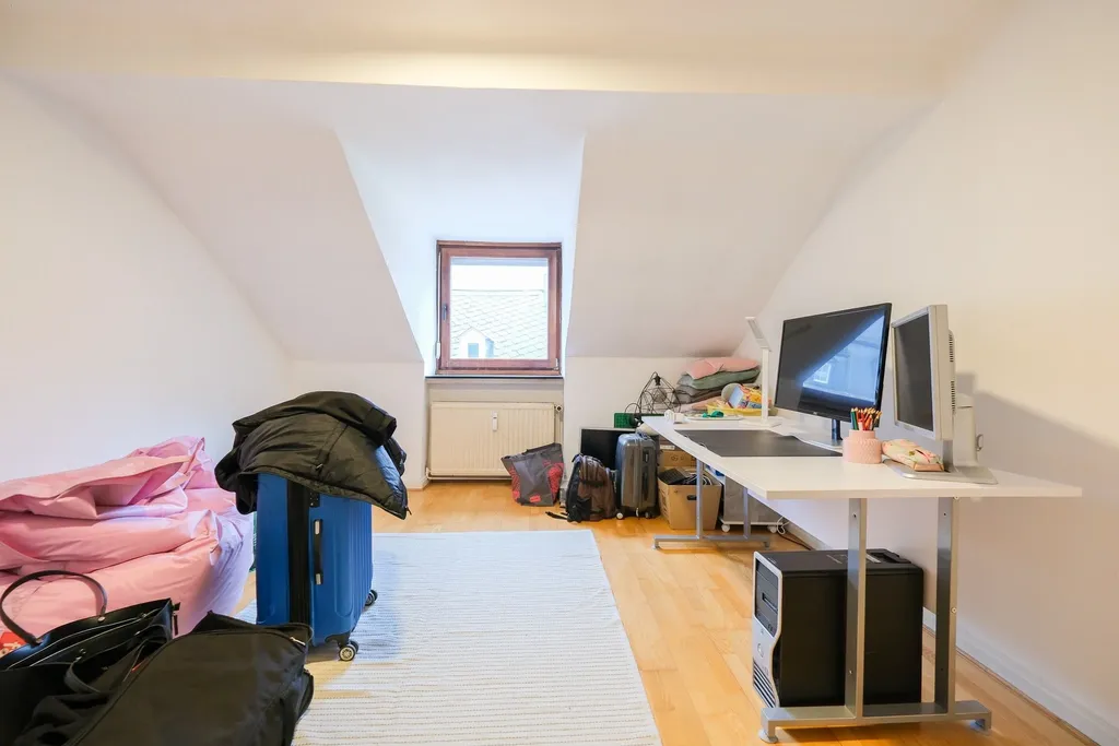 Bright attic room with sloping ceilings, window, and wood flooring, featuring a workspace with two monitors and PC tower, along with suitcases and boxes in the background.