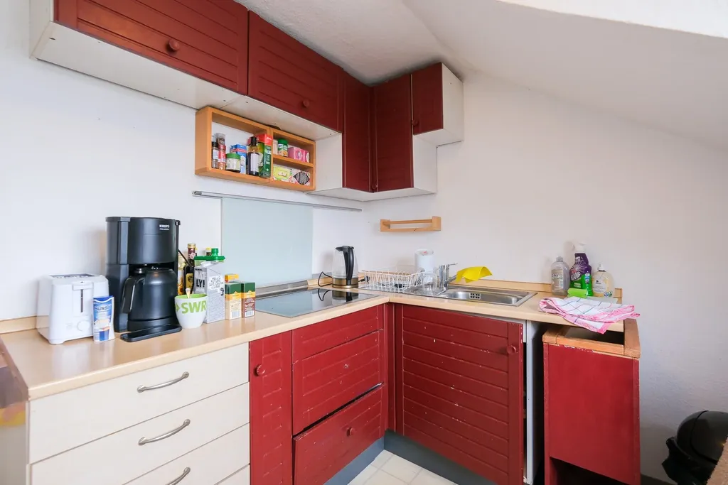 Compact modern kitchen with red cabinets, coffee maker, sink, and stovetop under a slanted ceiling.