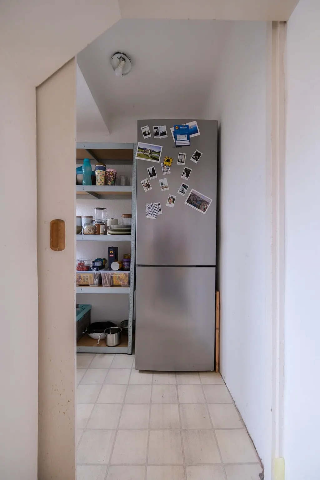 A stainless steel fridge freezer combination is placed in a white pantry next to shelves filled with jars, cans, and other kitchen utensils.
