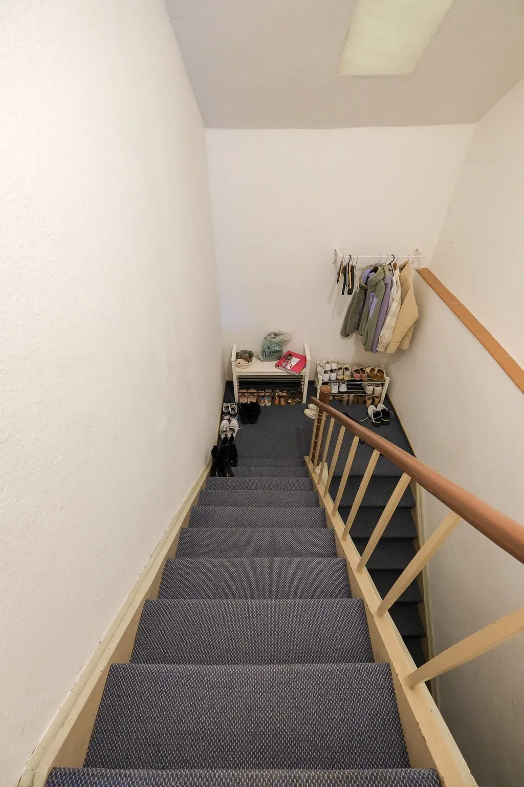 Staircase with blue-gray carpeting, shoe racks and coats hanging on a wall at the bottom, in an indoor setting.