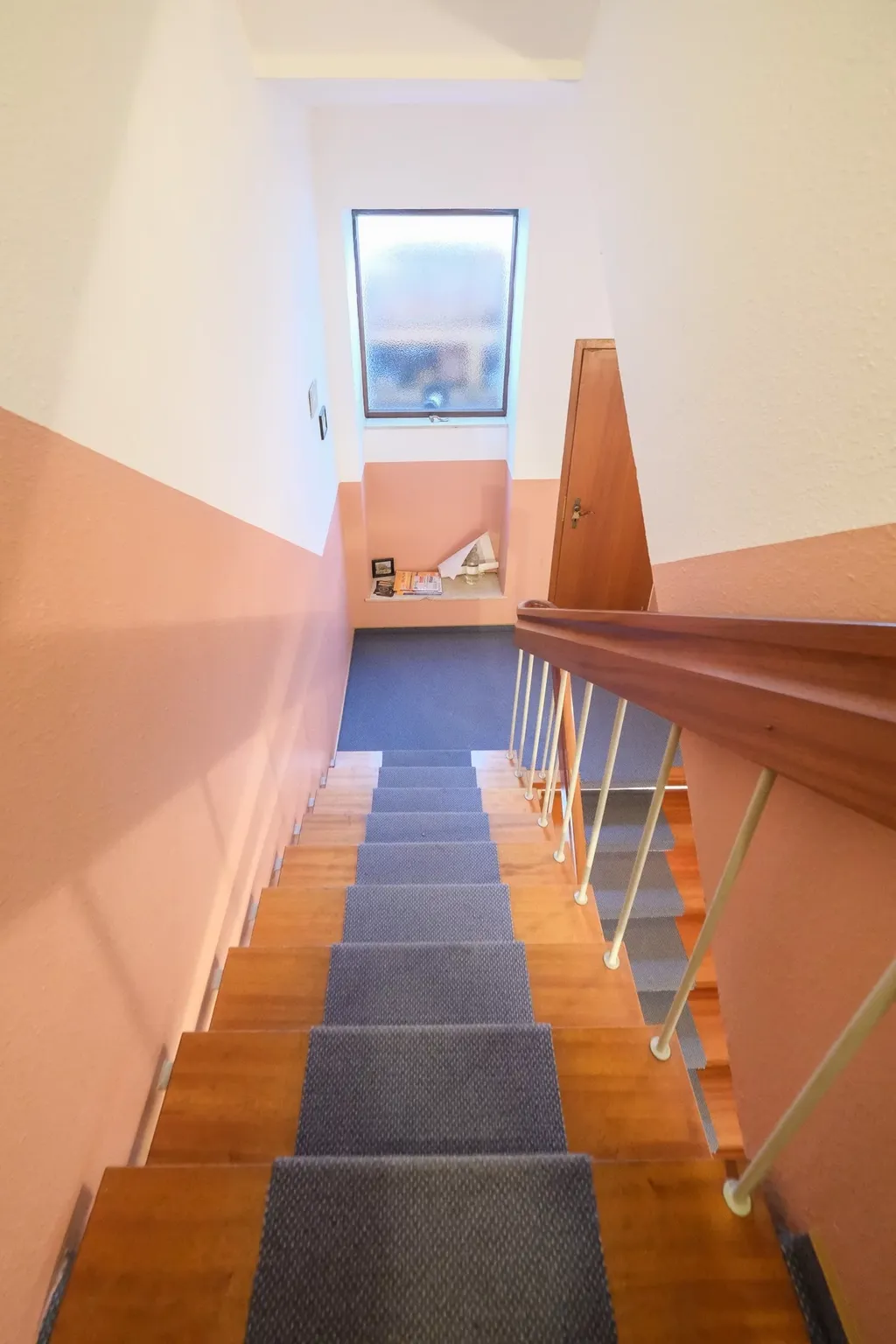 Staircase with dark blue carpet runner, beige walls, and a frosted glass window at the landing next to a wooden door.