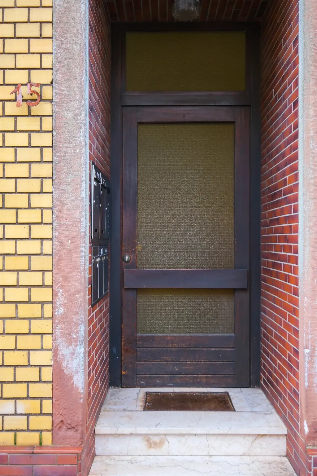 Entrance door of a building framed by yellow and red brick walls, house number 15 on the left wall, several mailboxes beside the door, two marble steps leading up to the dark door.