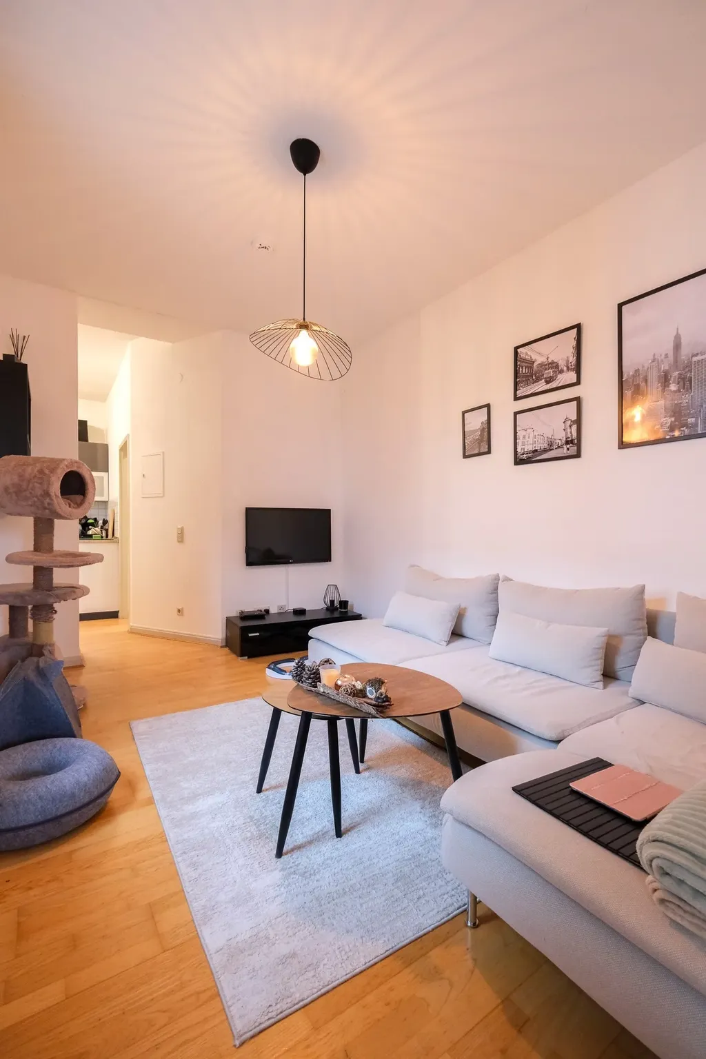 Bright living room with a large gray sectional sofa, two nested wooden coffee tables on a light rug, framed pictures on the wall, and a cat climbing tree beside the sofa.