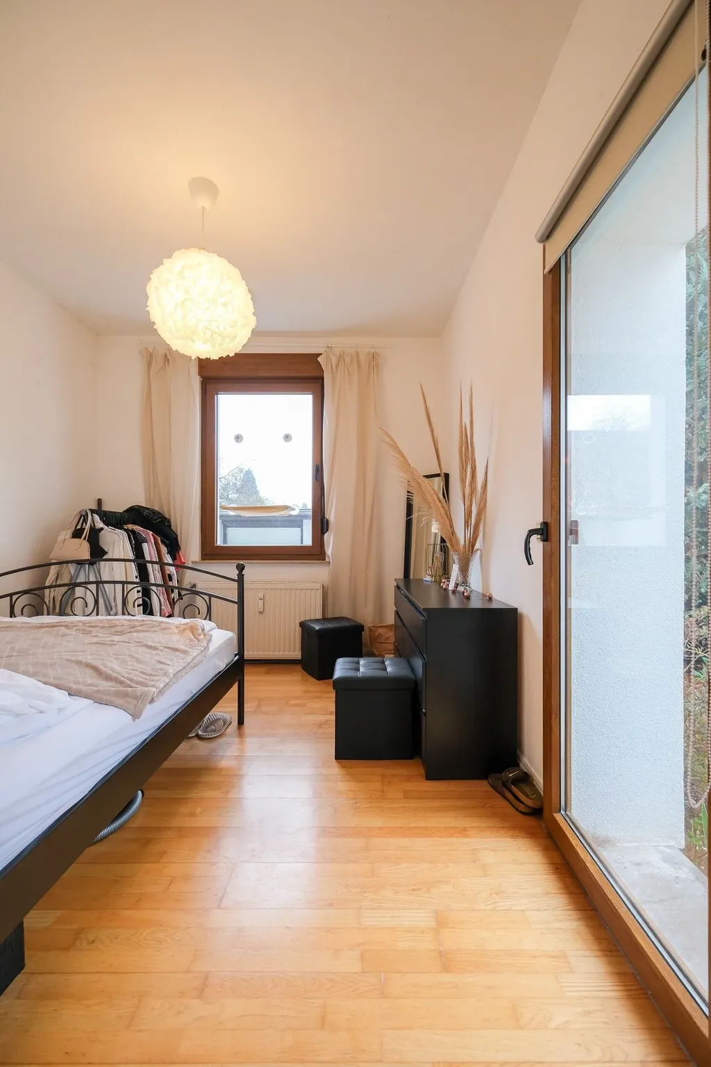 Brightly furnished bedroom with a metal bed, a window with beige curtains, a black dresser with decorative plants, and a large window overlooking a balcony.