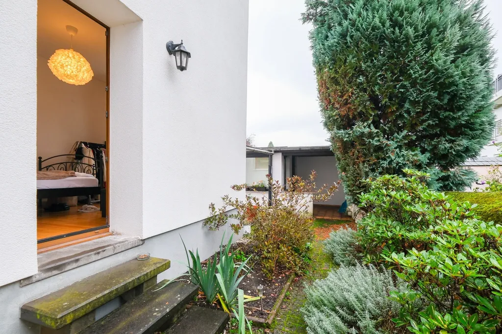 View of a garden with various plants and shrubs next to a white house with an open door revealing a bedroom with wooden flooring and a modern hanging lamp.
