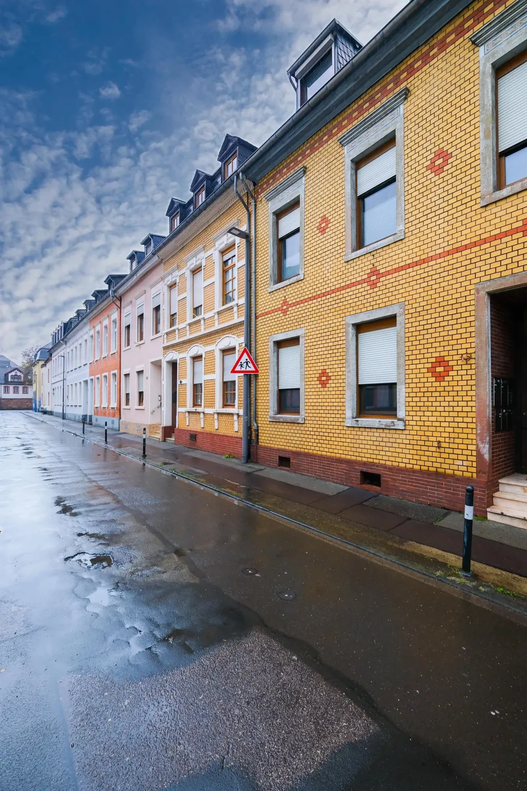 Row of colorful residential houses with yellow brick walls and white shutters along a wet street under a cloudy sky.