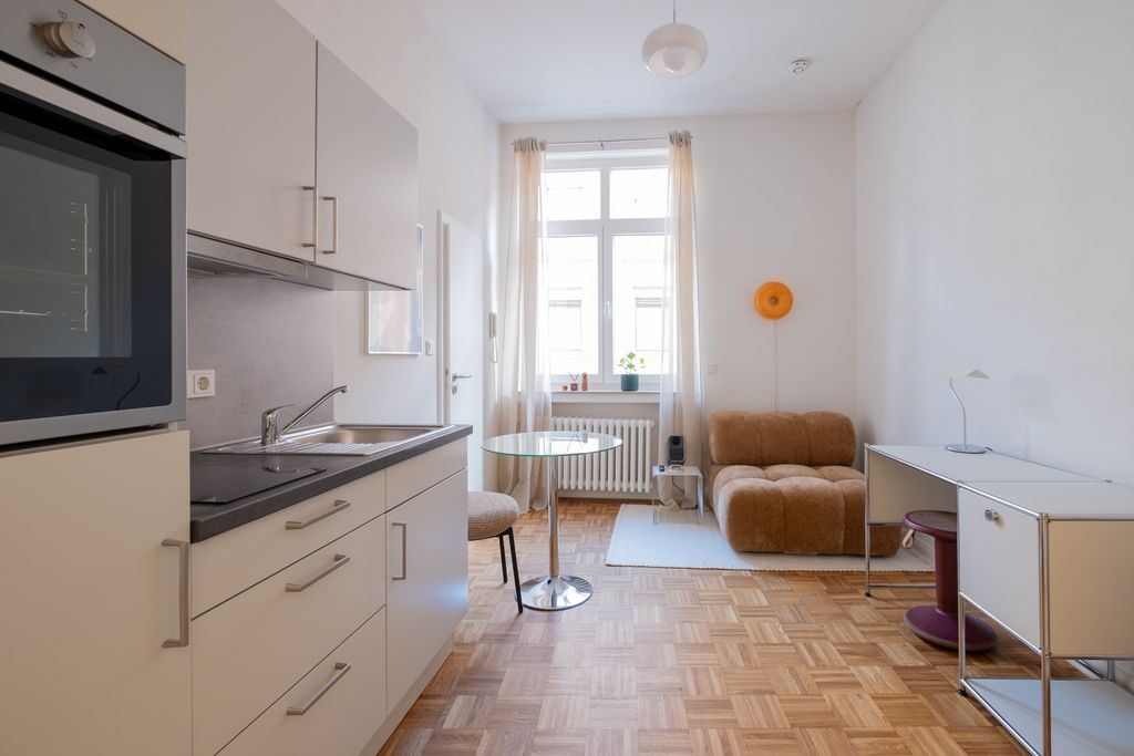 Modern bright studio apartment with kitchenette, glass dining area, brown armchair, and simple desk in front of a large window.