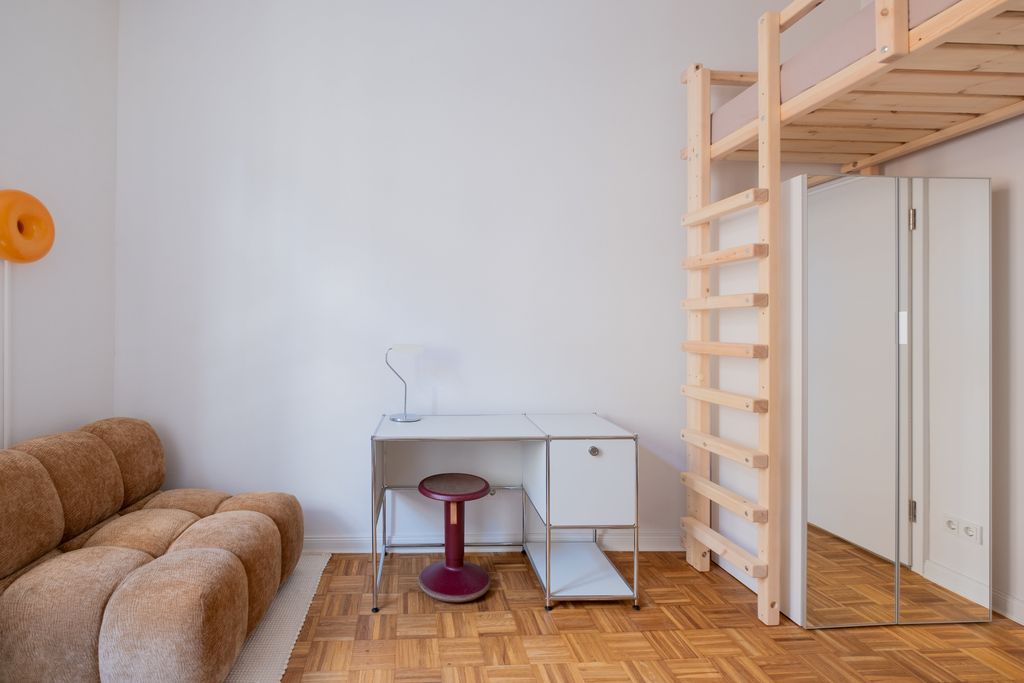 Minimalist room furnished with a brown sofa, white desk, red stool, and wooden loft bed with ladder next to a large mirror.