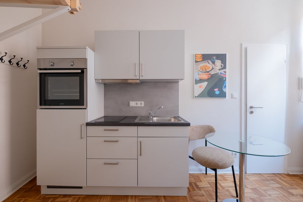 Small modern kitchenette with gray cabinets, built-in oven, stove, sink, and a glass table with two upholstered chairs in a bright room.