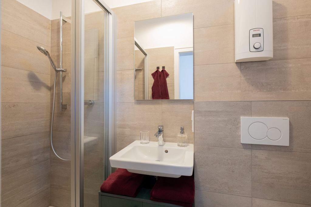 Small bathroom with shower cabin, white sink, mirror, and water heater mounted on the wall; red towels are visible under the sink.