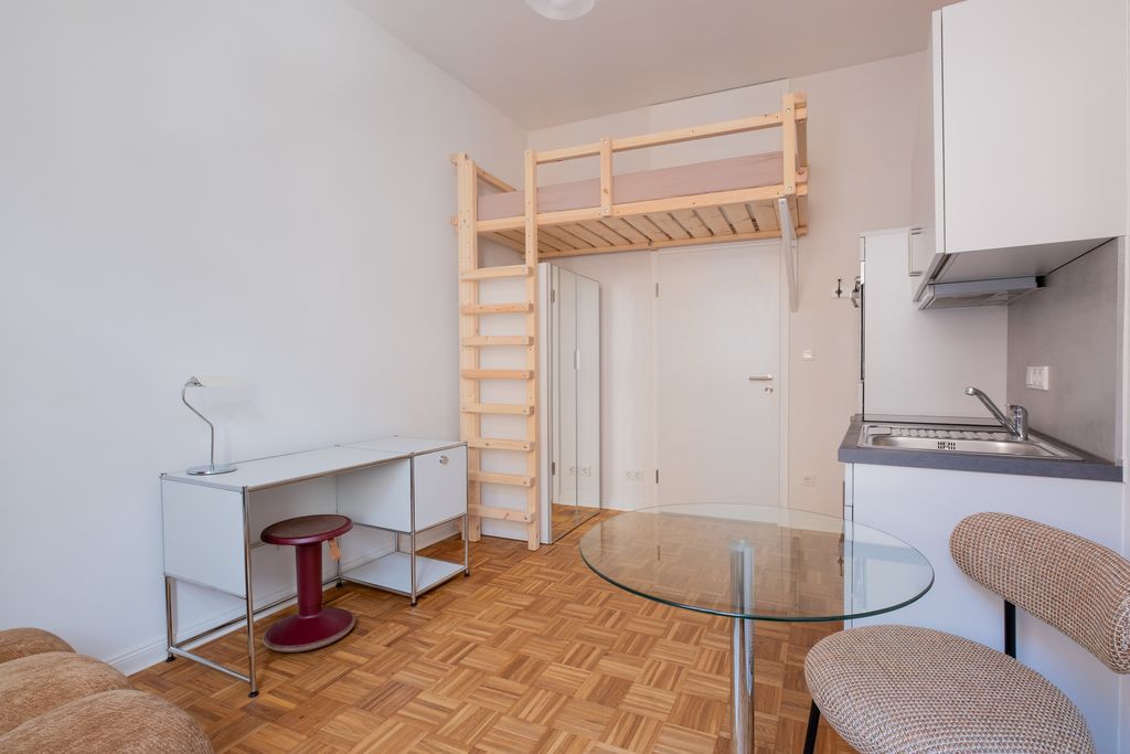 Small apartment with wooden flooring, white desk with red stool, elevated wooden bed, and kitchenette with sink and chair.