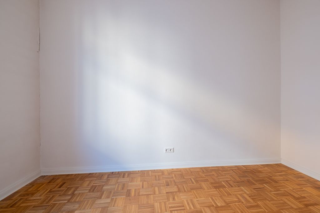 Empty room with light parquet flooring and white walls, sunlight casting on a wall with electrical outlets.