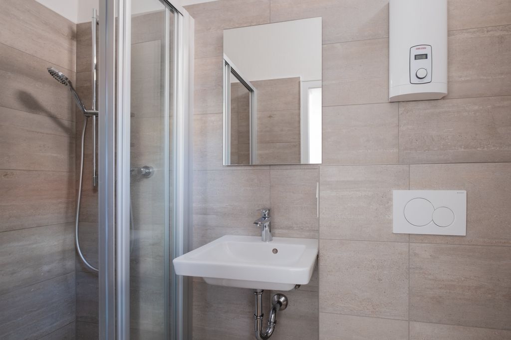 Small bathroom with shower, sink, mirror, wall heater, and wall-mounted toilet flush system on light tiled walls.
