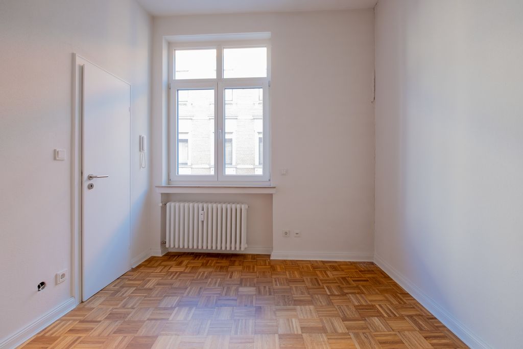 Empty room with white painted walls, a window overlooking a neighboring building, a radiator beneath the window, and a white door on the left side.