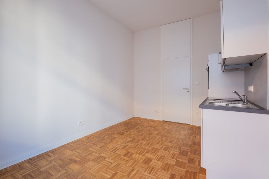 Empty room with white painted walls, wooden parquet flooring, and a small kitchenette with sink on the right side.