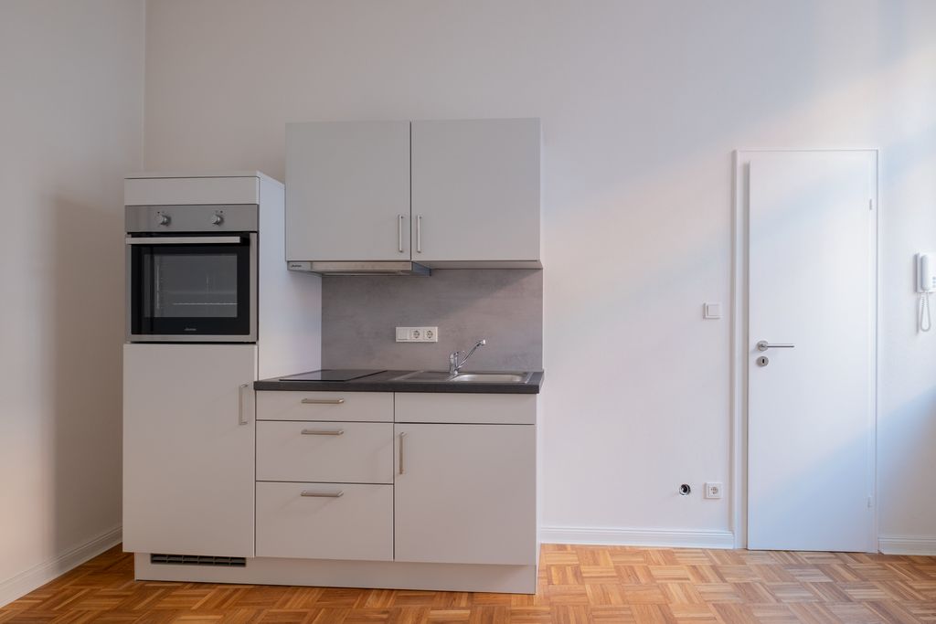 Small kitchenette in an empty, simply designed room with white walls, wooden floor, and a closed door to the right.