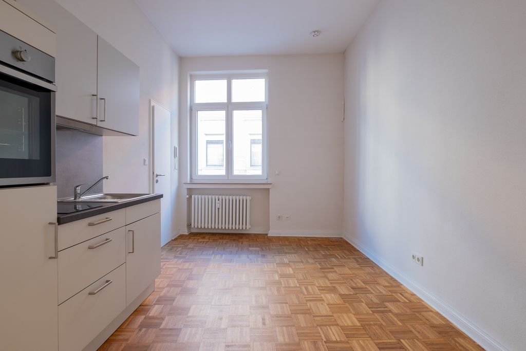 Empty, bright kitchen with white cabinets, oven and sink on the left, wooden floor and window with radiator on the back wall.