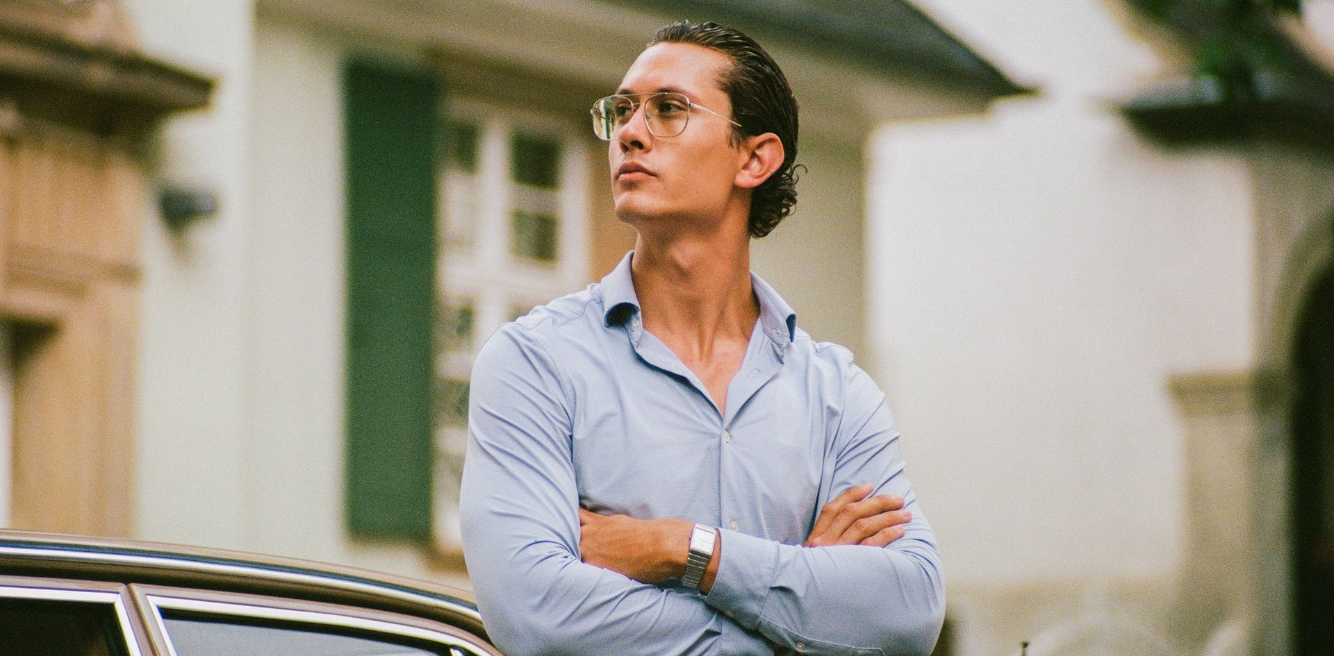 Junger Mann mit Brille und hellblauem Hemd steht mit verschränkten Armen neben einem Auto vor einem Gebäude mit grünen Fensterläden.