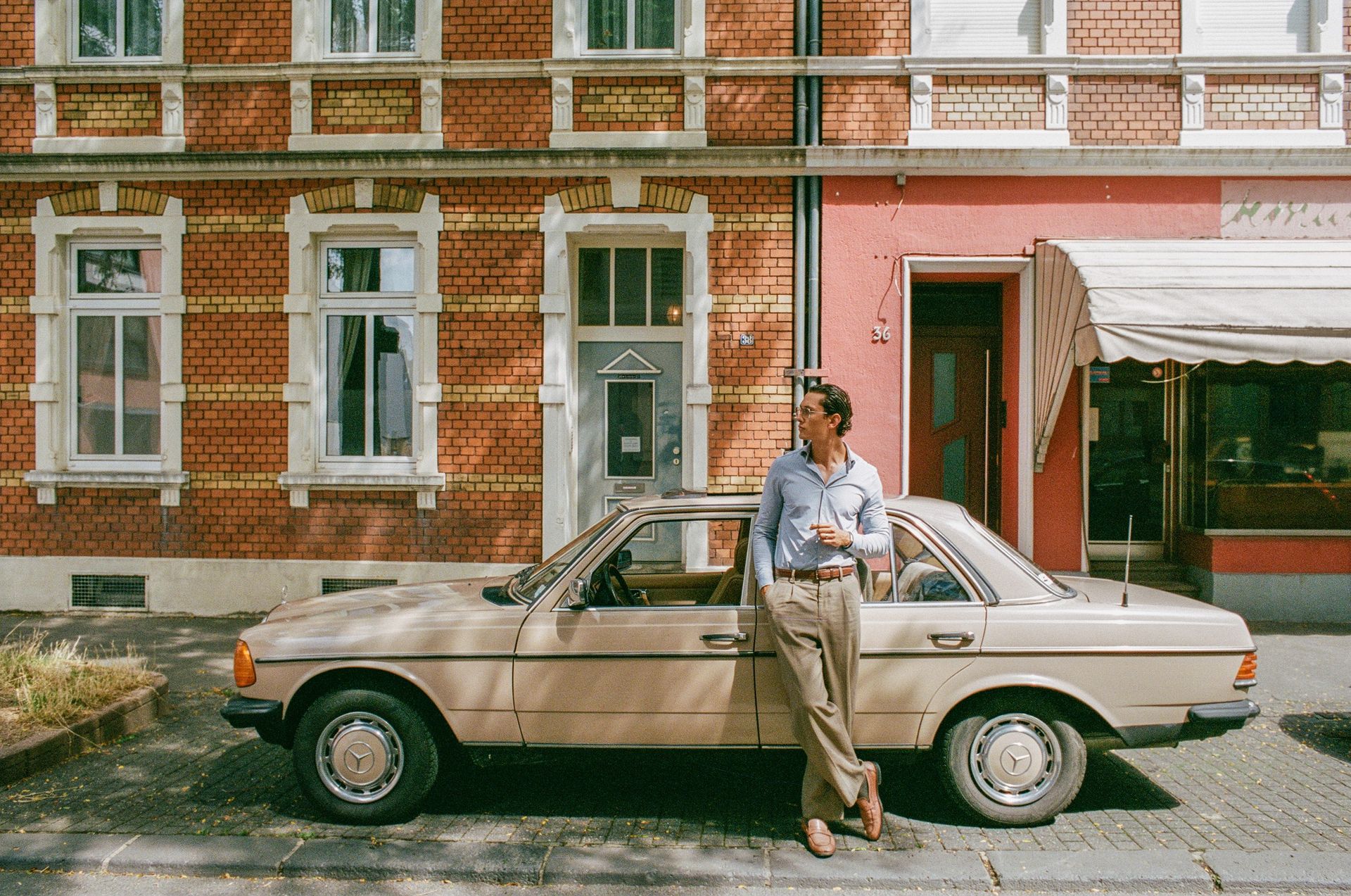 Ein Mann lehnt an einem geparkten, beigefarbenen Oldtimer-Auto vor einem Backsteinhaus mit weißen Fensterrahmen und einem roten Nebengebäude mit Markise.