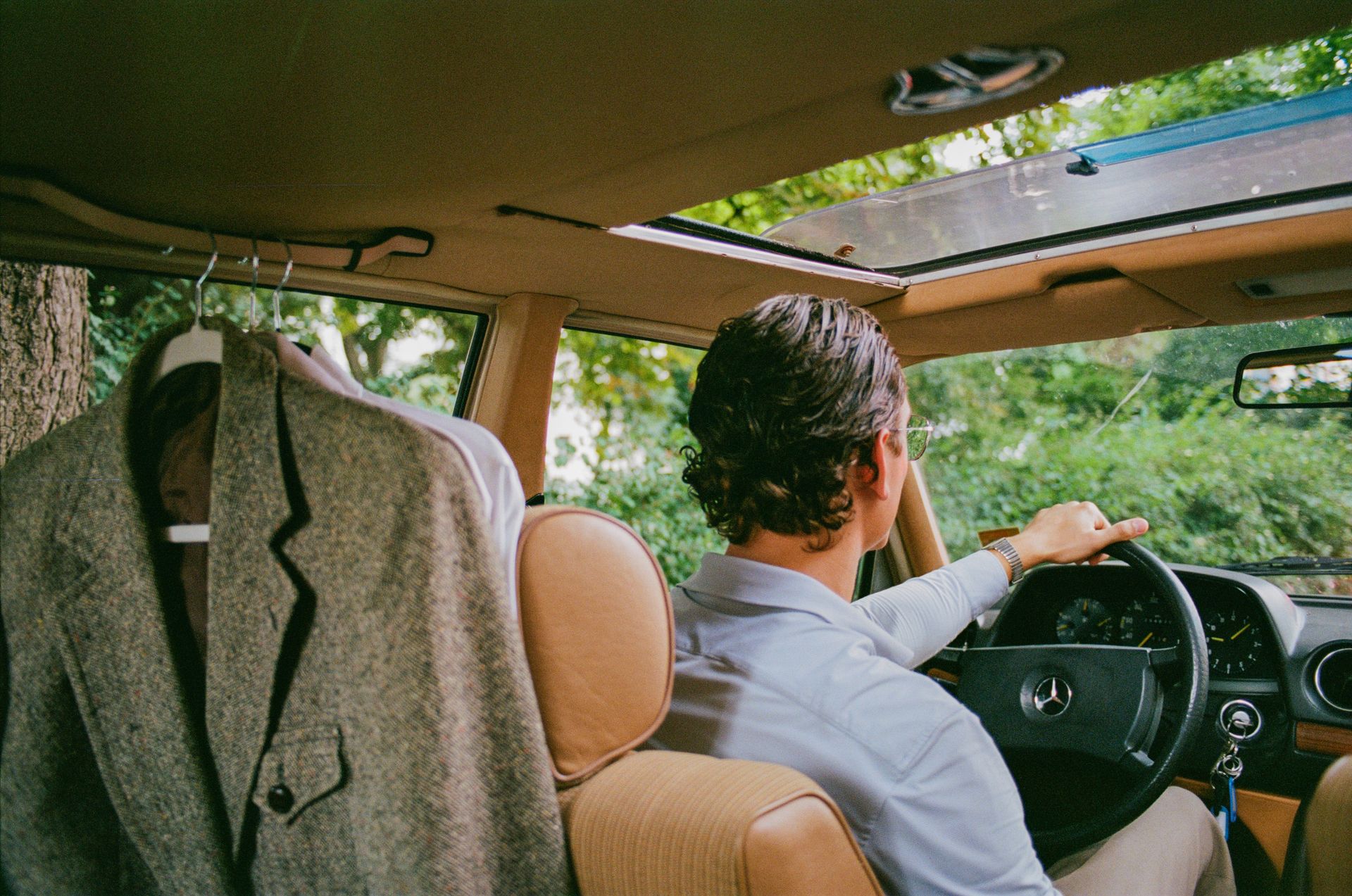 Ein Mann mit Hemd und Uhr lenkt ein Auto mit einem offenen Schiebedach, während ein grauer Anzug auf einem Kleiderbügel neben dem Fahrersitz hängt.