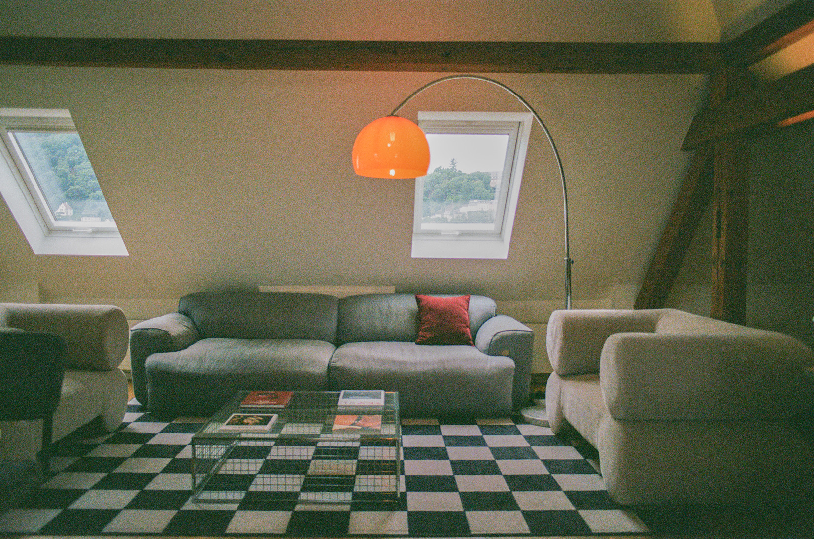 Ein stilvolles Wohnzimmer mit einem grauen Sofa, einem transparenten Couchtisch und einem orangefarbenen Bogenlampenschirm, aufgehellt durch zwei Dachfenster und mit schwarzem und weißem Schachbrettmusterteppich.