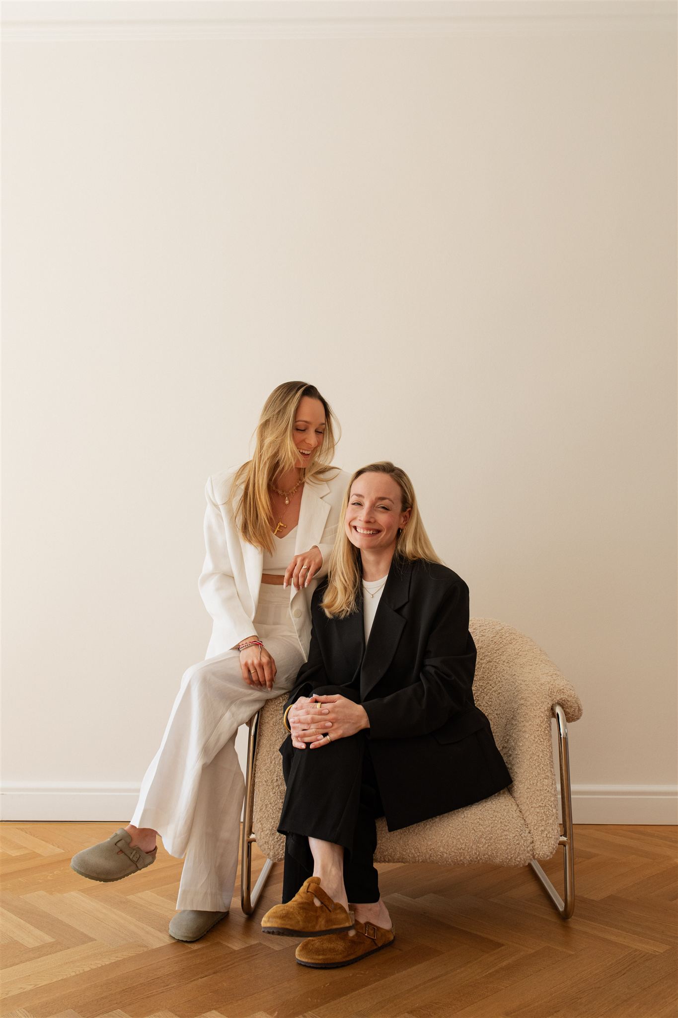Zwei lächelnde Frauen mit langen blonden Haaren, eine sitzt auf einem Becherstuhl in einem hellen Raum, die andere lehnt sich daneben. Beide tragen elegante Blazer und bequeme Hausschuhe.