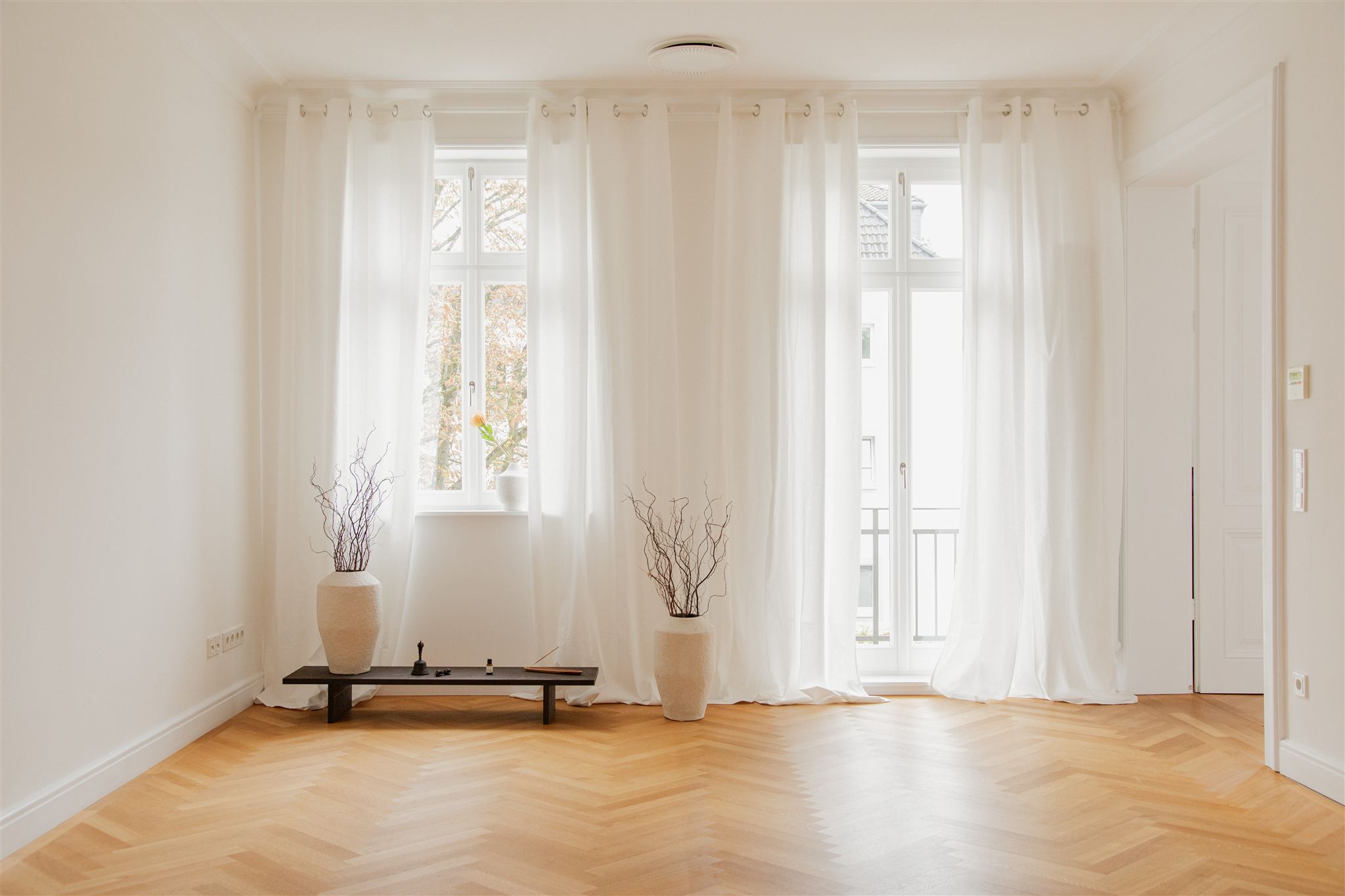 Ein heller, leerer Raum mit Holzboden und zwei großen Fenstern, die von weißen transparenten Vorhängen umgeben sind, auf dem Boden stehen zwei große beige Vasen mit dekorativen Zweigen.