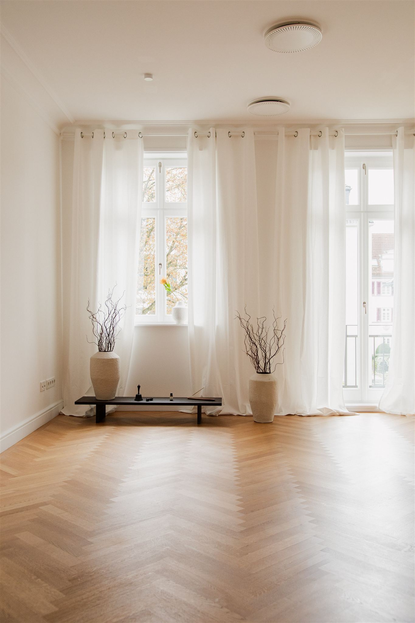 Helles, minimalistisch eingerichtetes Zimmer mit hellen Vorhängen vor zwei Fenstern, Holzparkettboden und zwei großen Vasen mit dekorativen Zweigen auf einem niedrigen schwarzen Tisch.