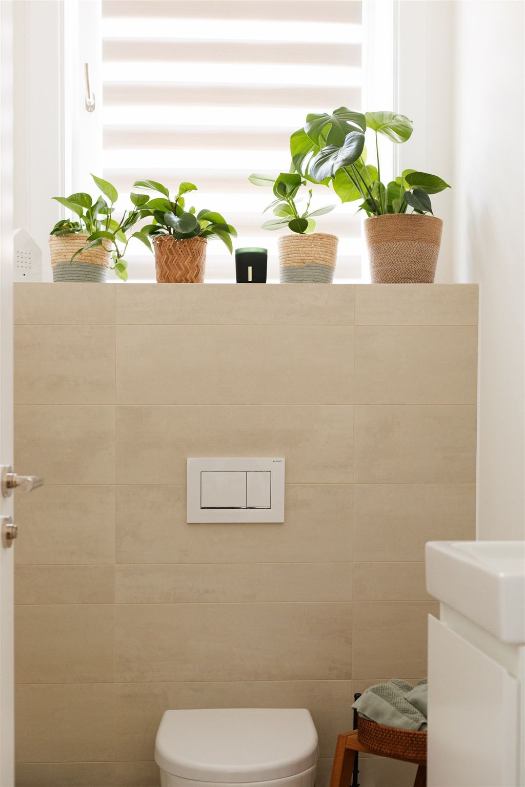 Ein minimalistisches Badezimmer mit beigen Wandfliesen, einer weißen Toilette und vier Topfpflanzen auf einer Fensterbank.