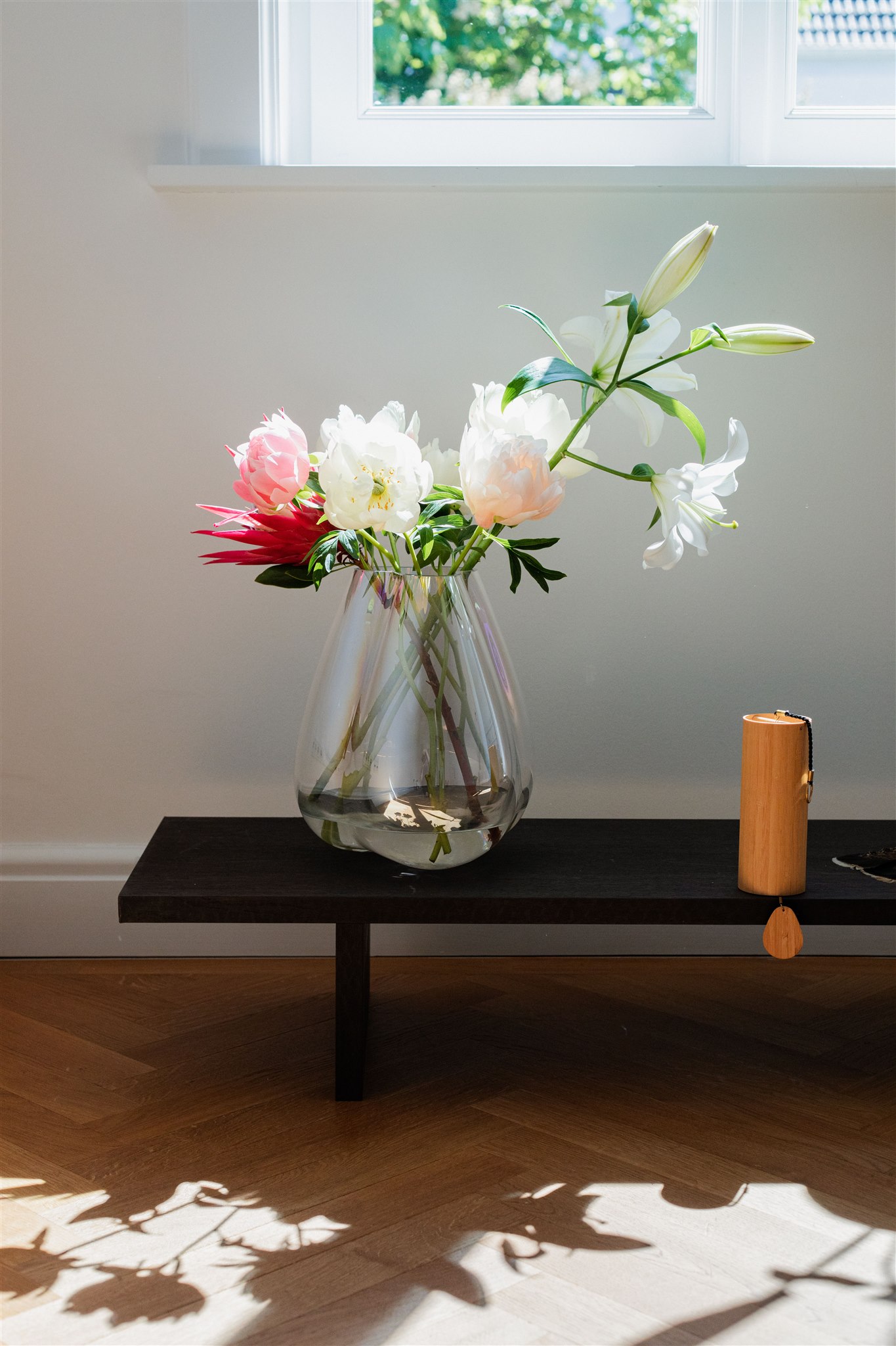 Ein transparenter Glasvase mit weißen und pinken Pfingstrosen und weißen Lilien auf einem dunklen Holztablett in der Nähe eines Fensters, mit Sonnenlicht, das Schatten auf den Holzfußboden wirft.