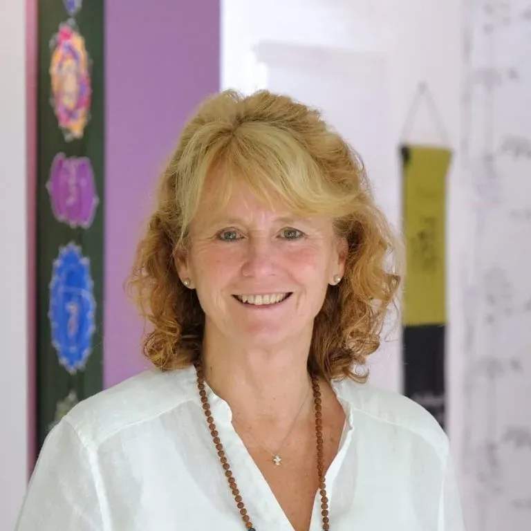 A smiling woman with ginger curly hair wearing a white shirt and a beaded necklace, standing in a colorful classroom or office setting.