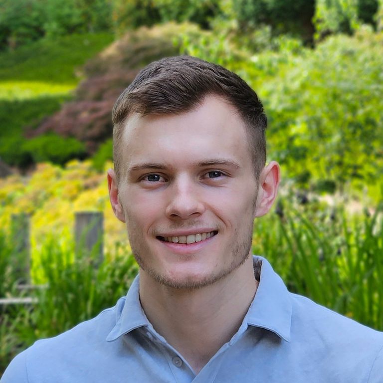 Portrait of a young man with short brown hair and blue shirt in front of a green, lush landscape.