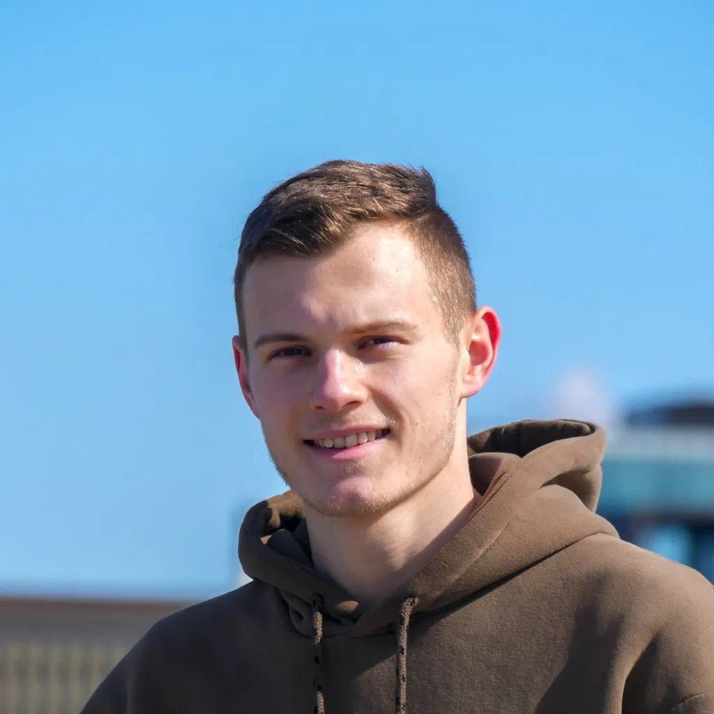 A young man with short brown hair and light skin, wearing a brown hoodie, standing outdoors against a clear blue sky.