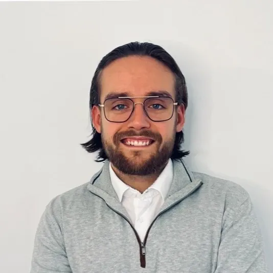 A young man with shoulder-length dark hair, glasses, and a beard, smiling and wearing a light gray zip-up sweater over a white shirt against a plain white background.