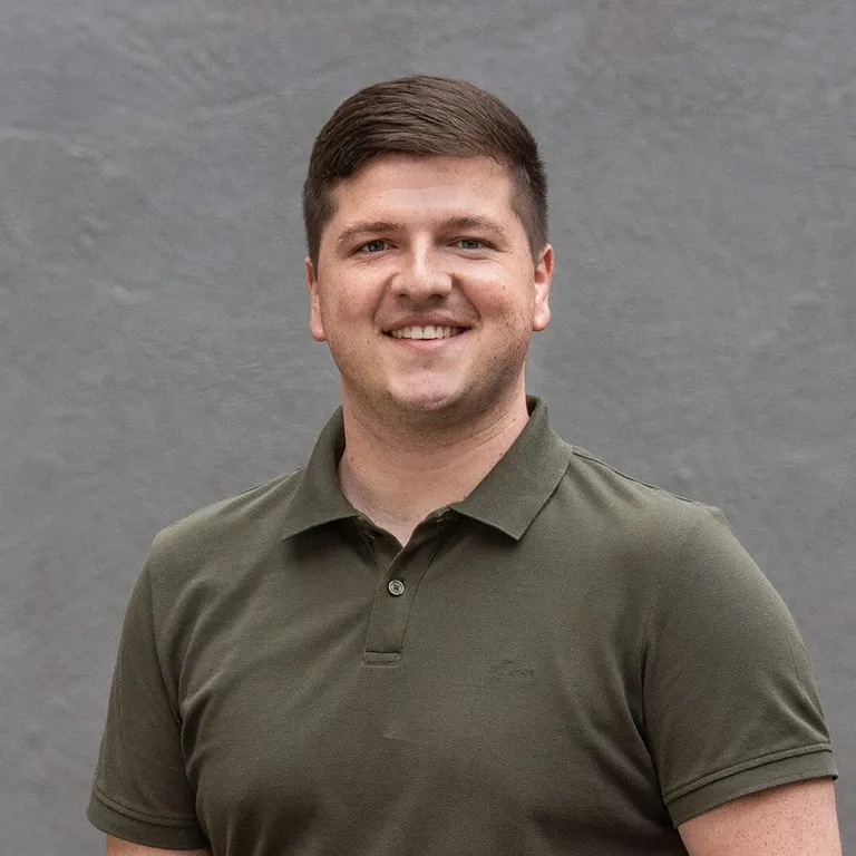 A smiling young man with short brown hair wearing a green polo shirt standing outdoors against a gray wall.