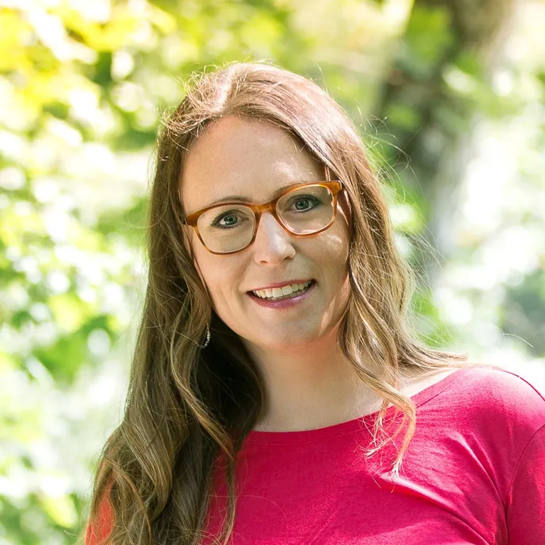 A woman with long light brown hair and glasses wearing a red top standing outdoors with greenery in the background.