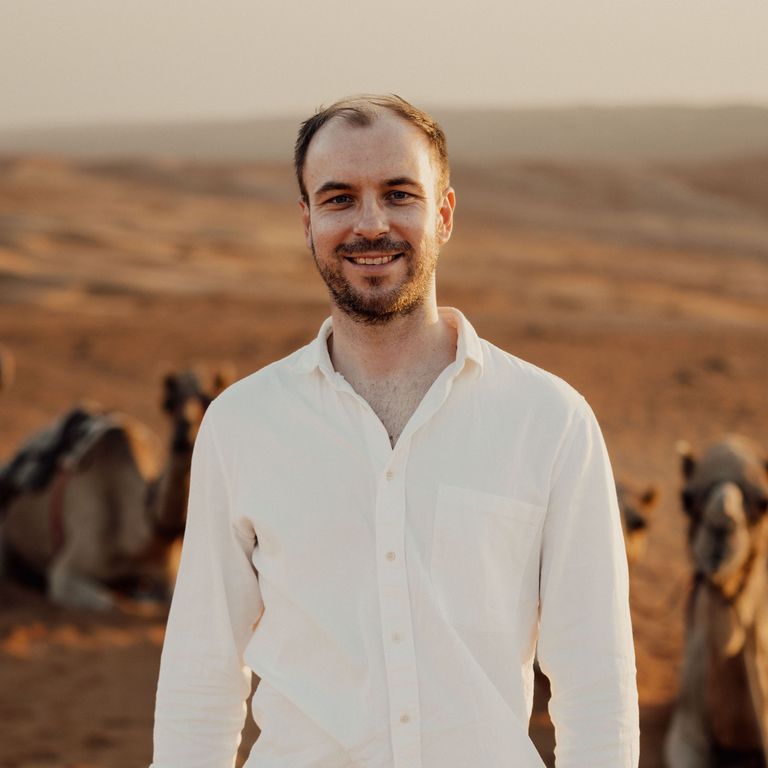 A smiling man with short brown hair and a beard, wearing a white shirt, standing outdoors with camels in the background during sunset.