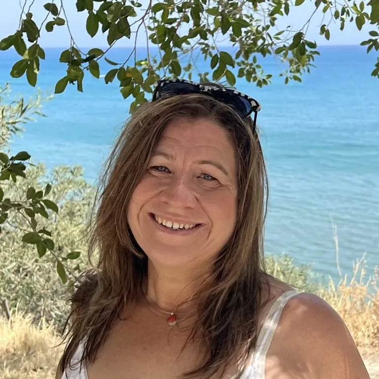 A woman with long brown hair smiling outdoors near the sea with leafy branches above.