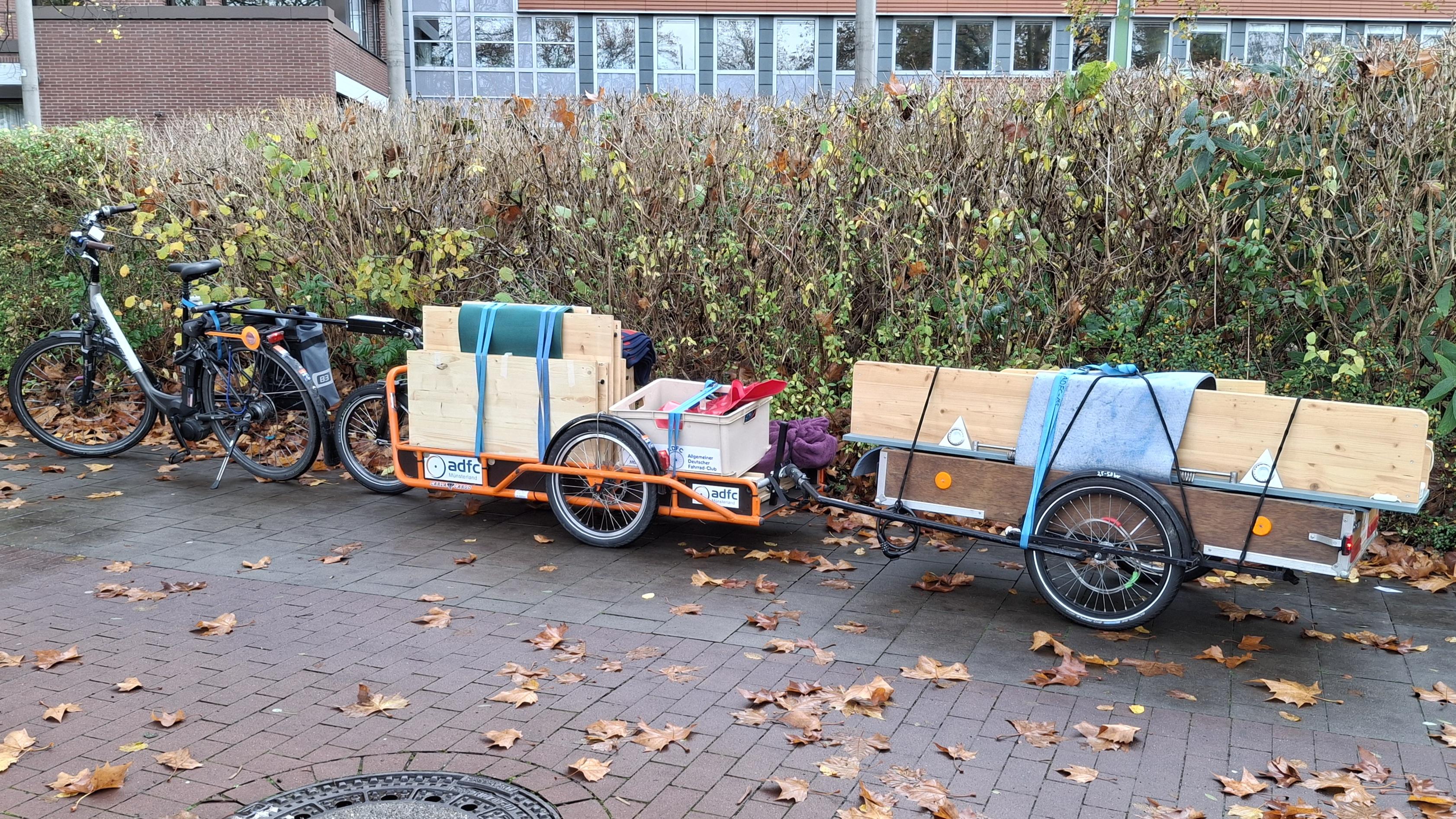 Vor einer Hecke steht ein Fahrrad-Lastenanhängergespann, bestehend aus 2 Lastenanhängern. Der erste Anhänger ist eine CarlaCargo mit Auflaufbremse, die mit den Teilen von 3 Unterschränken sowie kleinteilen in einer Kunststoffkiste beladen ist. Der zweite Anhänger ist an die CarlaCargo angekoppelt auf ihm liegen die Hängeregale.