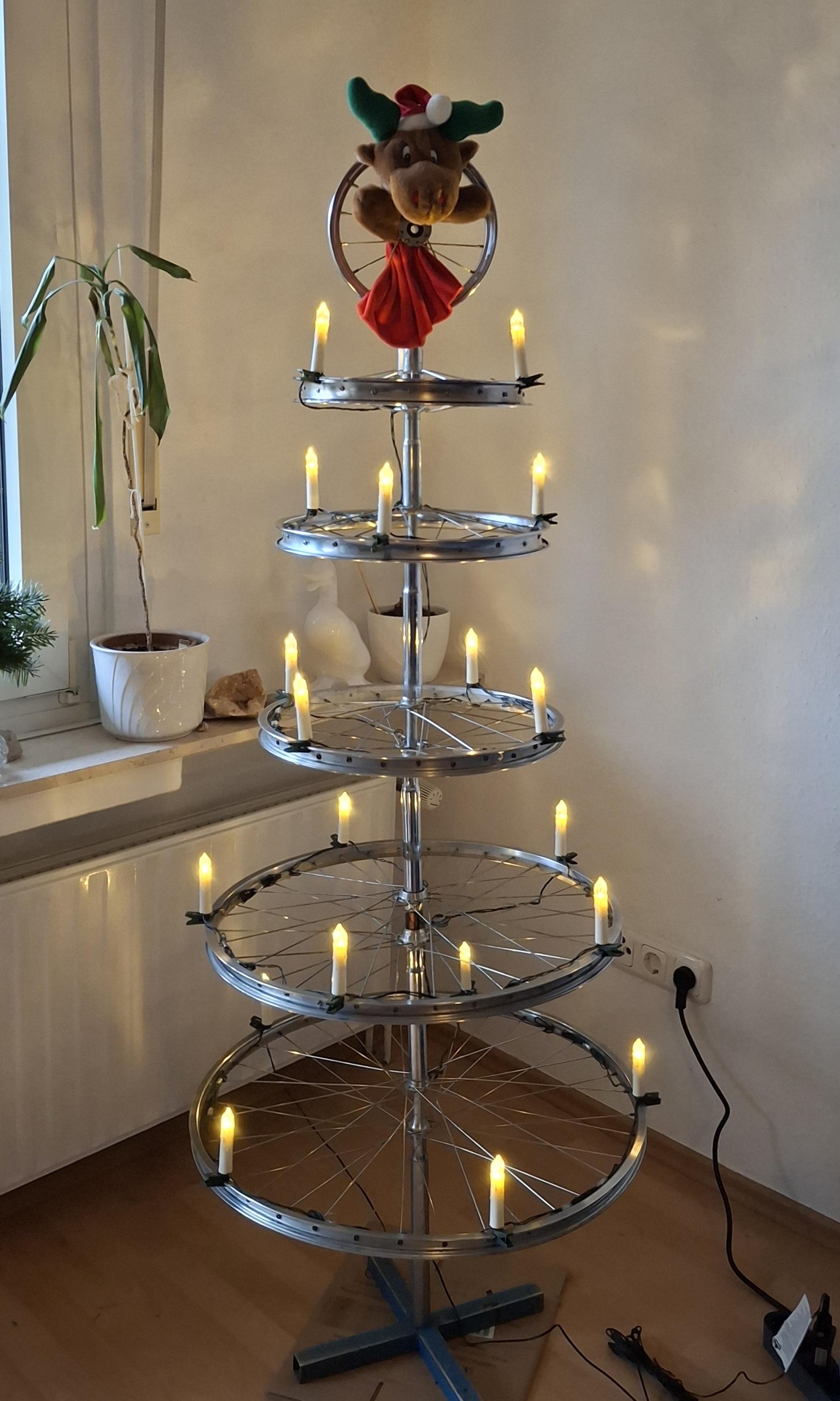 In einer Wohnzimmerecke vor der Fensterbank steht ein aus Fahrrafvorderrädern und Sattelstützen zusammen gebauter Weihnachtsbaum. Auf den blanken Felgen der Laufräder sind 20 LED-Kerzen verteilt. Oben aus der senkrecht stehenden 12" Felge schaut eine Rudolfhandpuppe heraus.