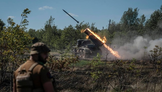 Military truck fires a large rocket in a rural field, flames and smoke trailing from the launcher while a soldier watches in the foreground.