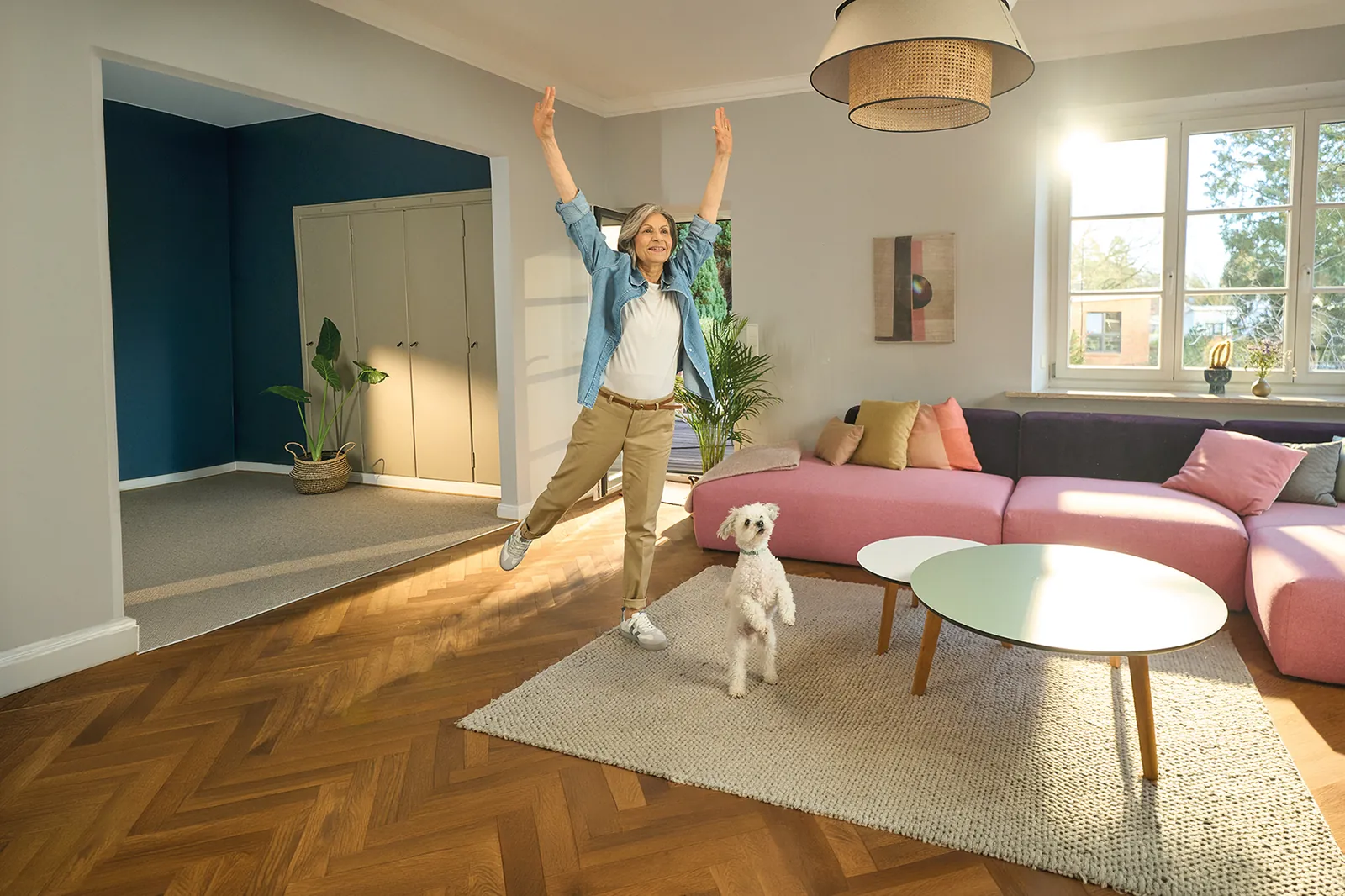 Lebensfrohe Frau macht Dehnübungen im Wohnzimmer, während ihr kleiner weißer Hund spielerisch neben ihr steht.