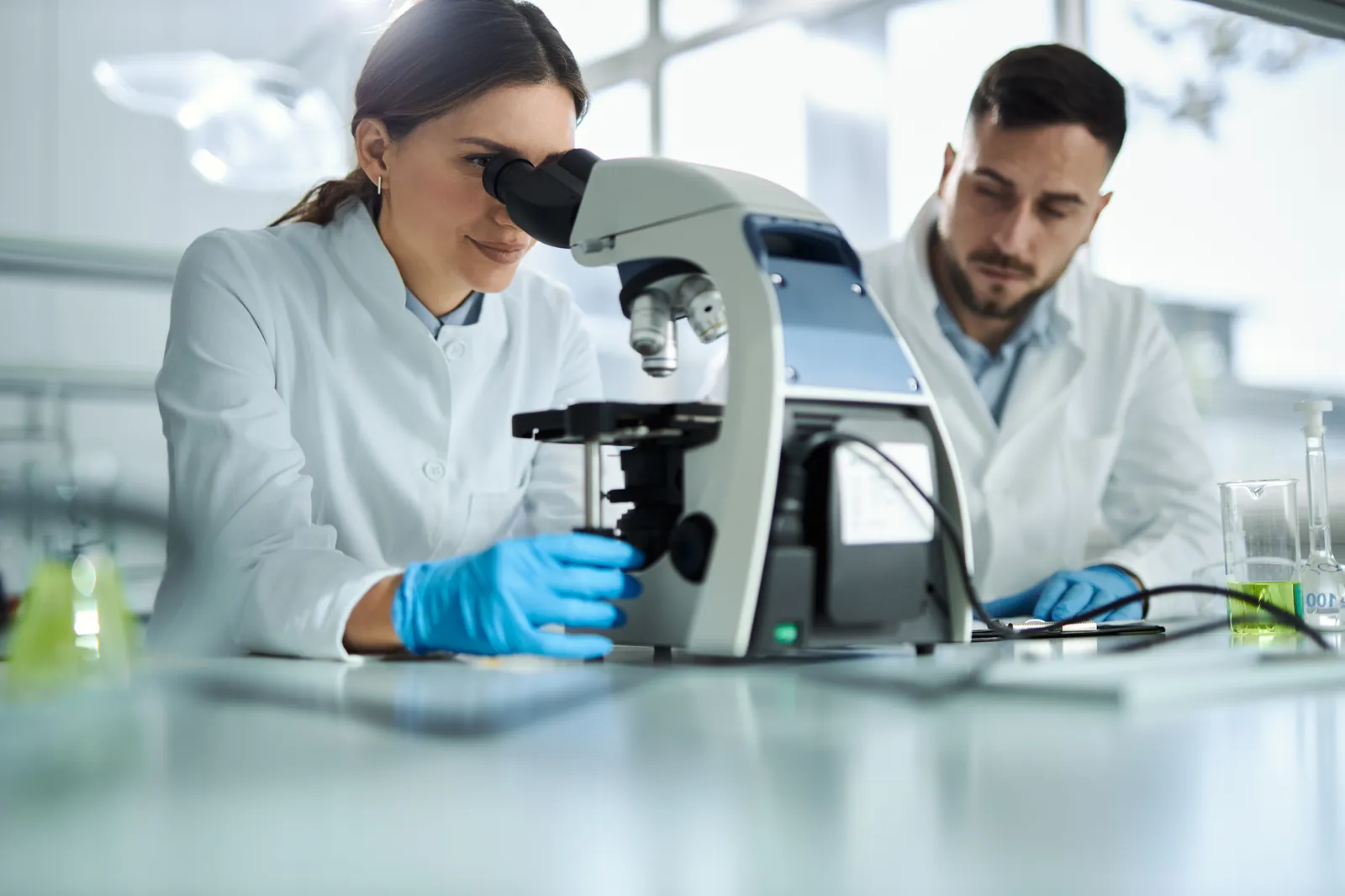 Zwei Wissenschaftler im weißen Laborkittel arbeiten im Labor; eine Frau blickt konzentriert durch ein Mikroskop.