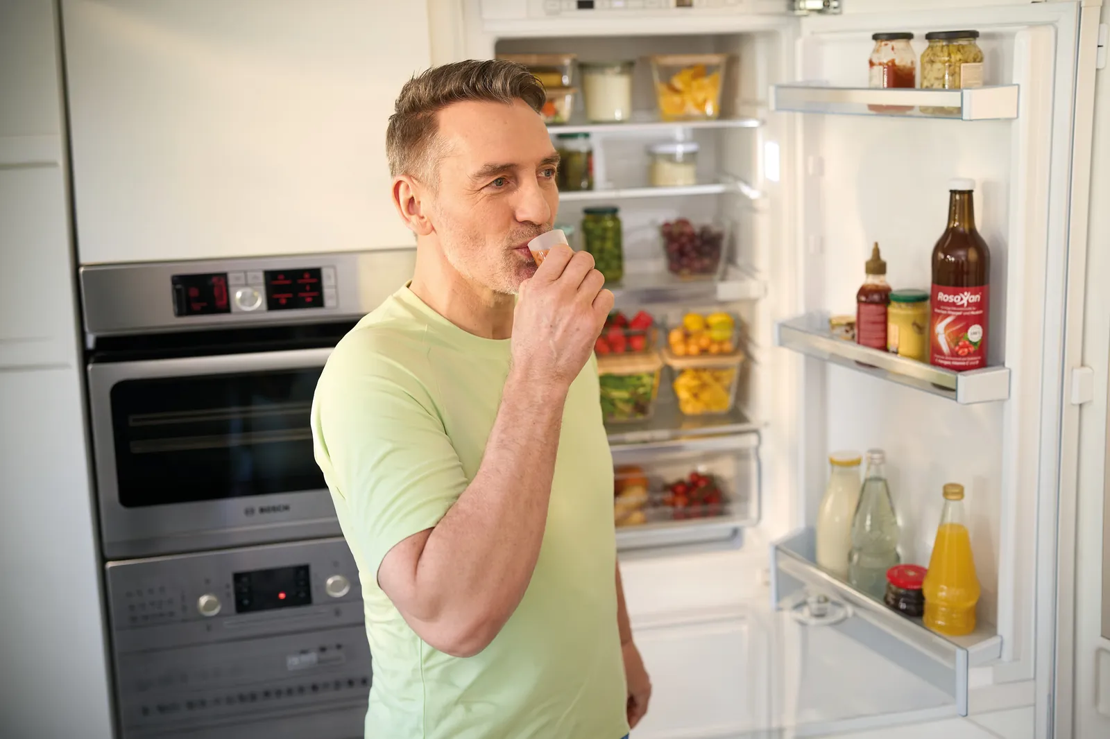 Ein Mann in einer Küche trinkt eine Portion Rosaxan aus einem Messbecher im geöffneten Kühlschrank steht eine Flasche Rosaxan