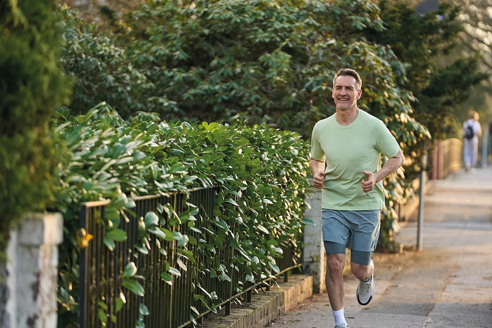 Ein Mann joggt lächelnd auf einem Gehweg im Freien an einer grünen Hecke entlang.