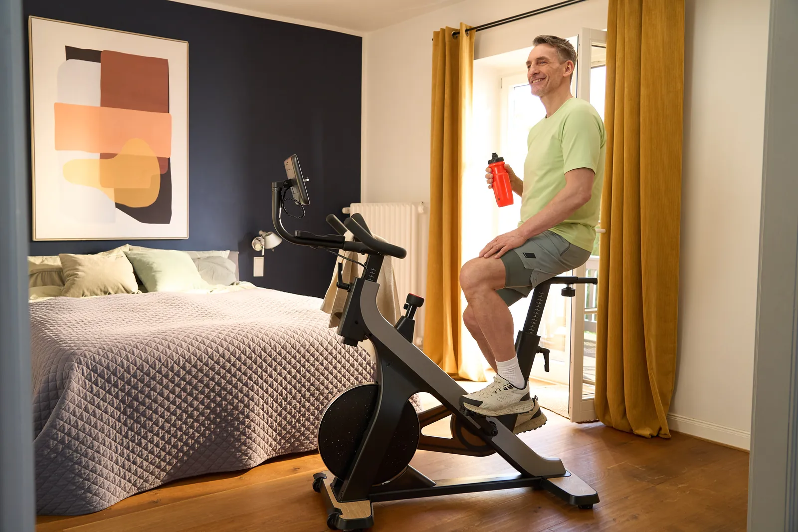 Ein lächelnder Mann sitzt entspannt auf einem Heimtrainer im Schlafzimmer und hält eine rote Trinkflasche in der Hand.