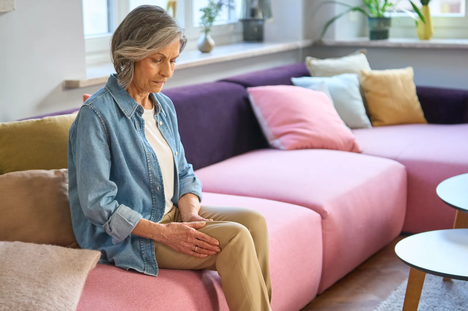 Eine ältere Frau sitzt besorgt auf einem Sofa und hält sich mit beiden Händen ihr schmerzendes Knie.