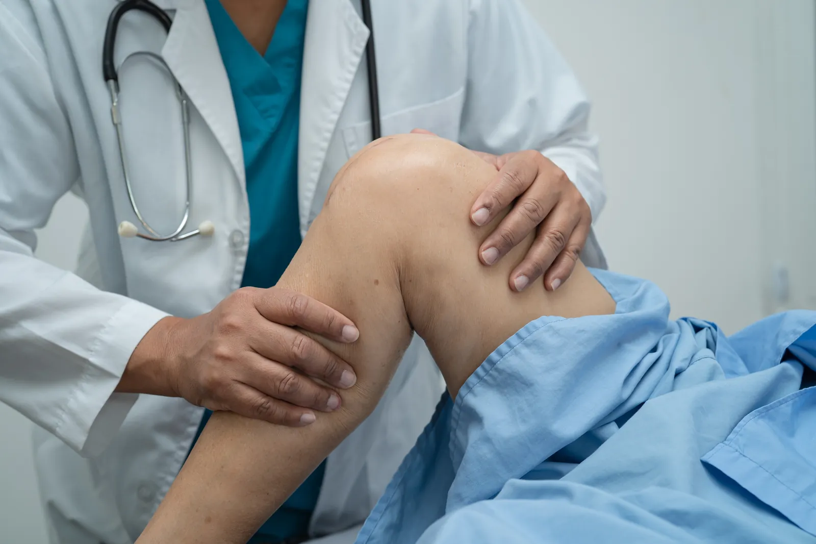 Nahaufnahme eines Arztes in weißem Kittel, der beide Hände zur Untersuchung an das angewinkelte Knie eines Patienten legt.