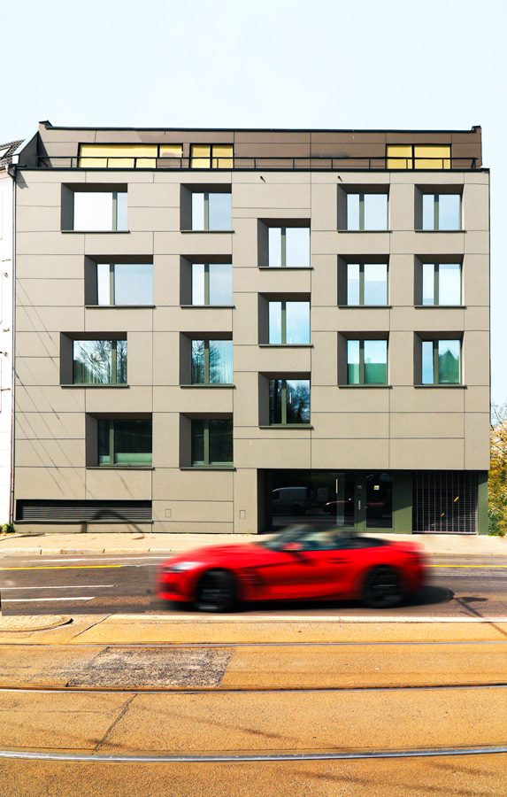 Frontale Straßenfassade des Neubaus mit grauen Faserzementplatten, unterschiedlich angeordneten Fenstern und zurückgesetztem Erdgeschoss; im Vordergrund Fahrbahn mit Straßenbahngleisen und ein vorbeifahrendes rotes Auto.